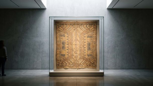 Vitrina de museo con un tejido ritual utilizado para cubrir una entrada, iluminada desde arriba en una tranquila sala de exposición.