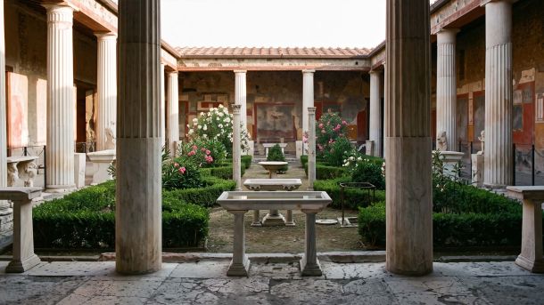 Jardín peristilo romano iluminado por el sol con columnata de mármol, setos cuidadosamente recortados y una fuente central, el tipo de jardín cerrado que las figuras guardianas grecorromanas protegían.