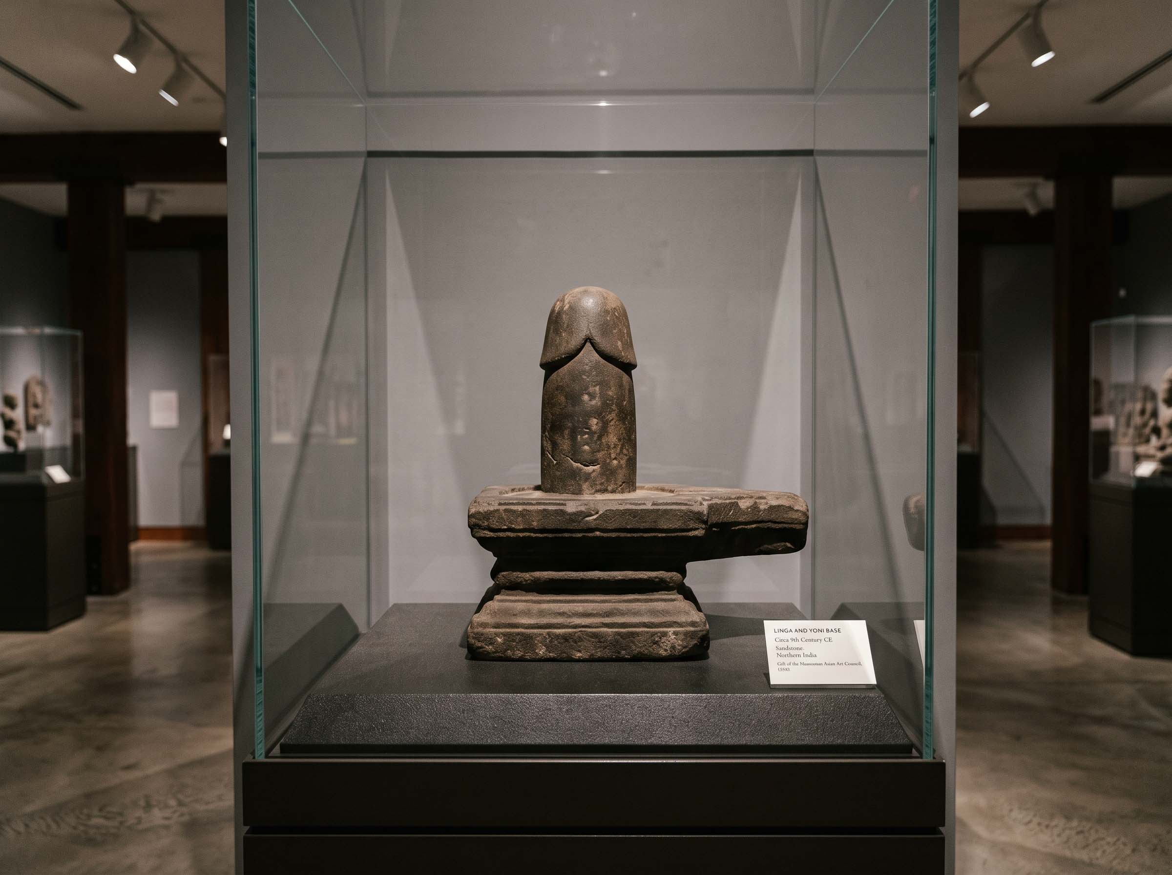 An early carved stone linga mounted on a grooved pedestal, displayed in a museum vitrine with archival lighting