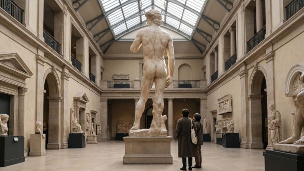 Visitors viewing a towering plaster nude sculpture in a museum cast court, illustrating cultural rules about the exposed body