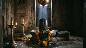 A polished stone Shiva lingam in its grooved pedestal inside a temple sanctum, surrounded by marigold offerings during daily ritual worship
